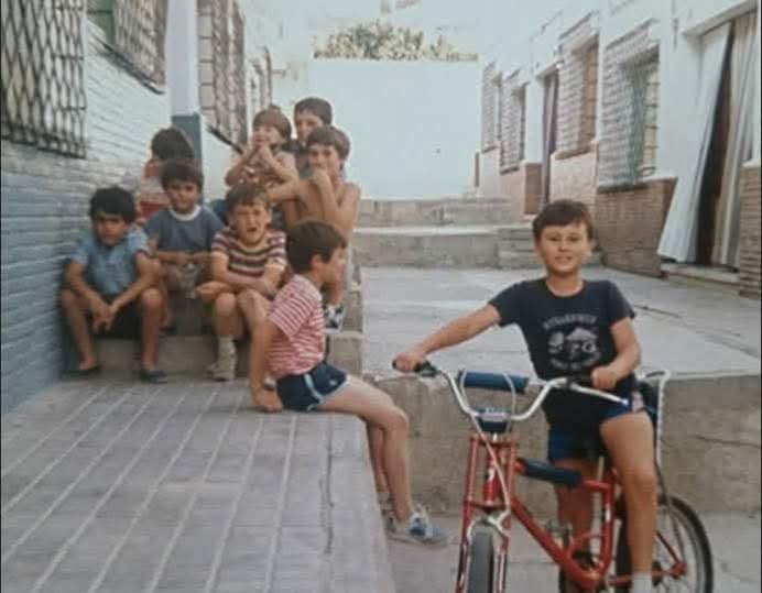 Grupo de niños sentados en la acera y un niño en bicicleta en una calle de pueblo