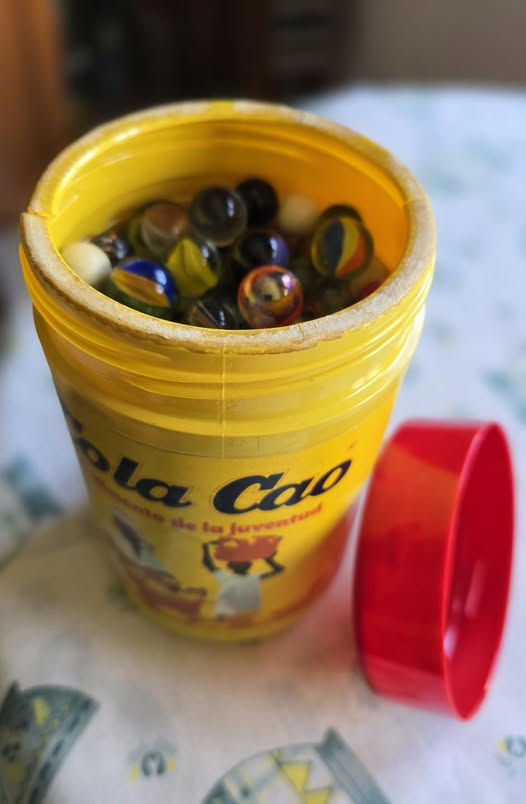 Bote amarillo de Cola Cao lleno de canicas con la tapa roja al lado.
