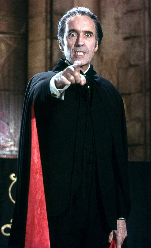 Christopher Lee como Drácula, con capa negra forrada de rojo, colmillos y señalando con el dedo.