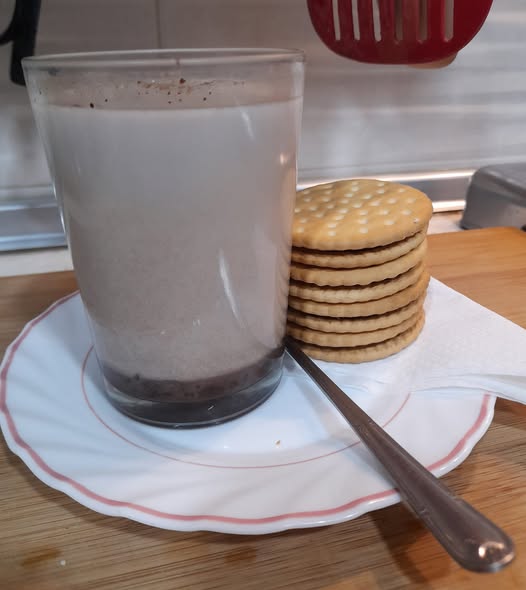 Vaso de leche con cacao junto a un montón de galletas y una cuchara.