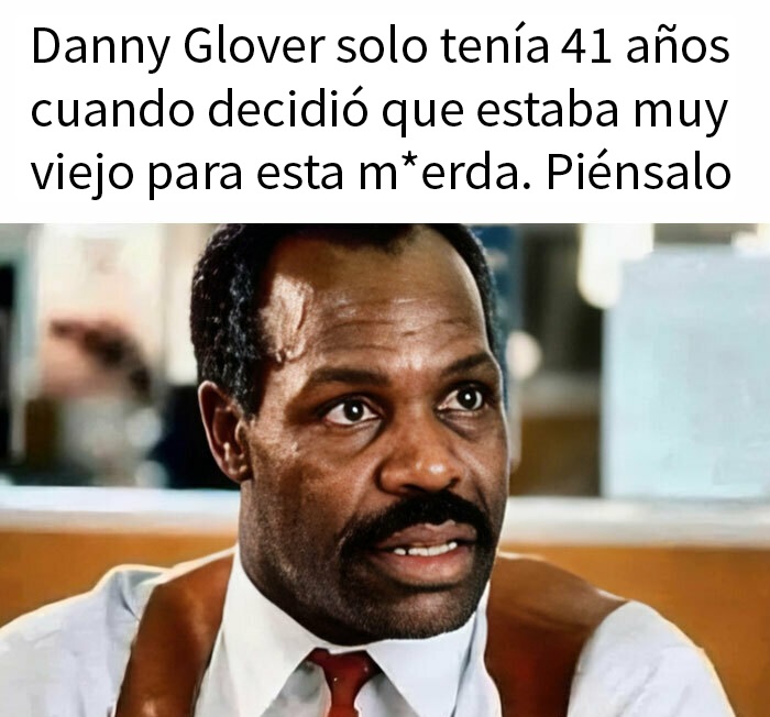 Danny Glover, con bigote, camisa blanca, chaleco y corbata roja, en un fotograma de Arma letal.