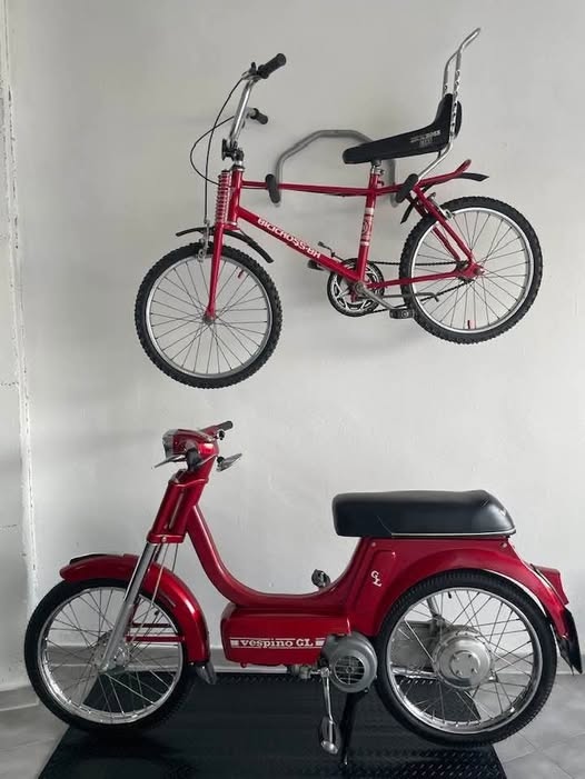 Bicicleta roja colgada de la pared y una moto Vespino roja debajo.