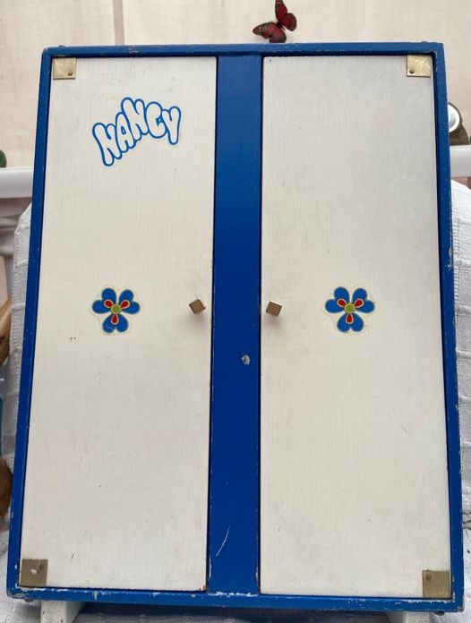Armario de juguete de madera blanco con bordes azules, dos puertas, el logotipo de Nancy y dos flores.