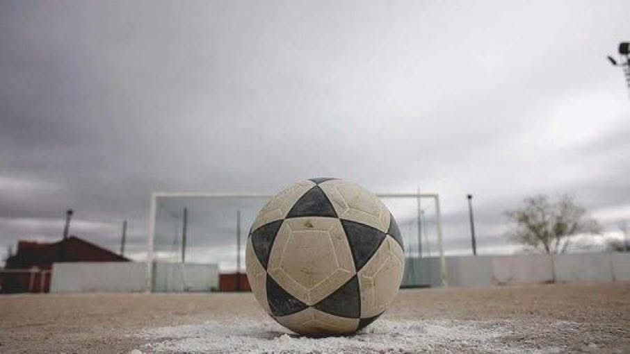 Balón de fútbol viejo sobre la cal de un campo de tierra, con una portería al fondo y cielo nublado.