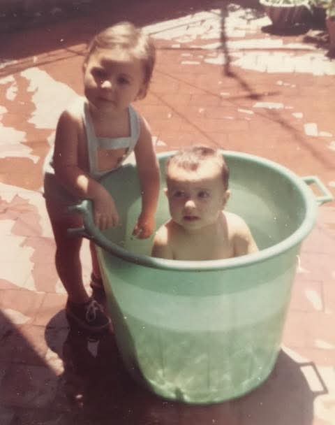 Foto antigua de un bebé bañándose en un barreño verde en una terraza mientras otro niño se asoma.