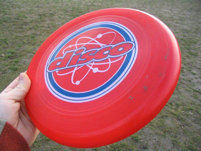 Mano sujetando un disco volador de plástico rojo con la palabra disco en el centro, sobre un campo de hierba.