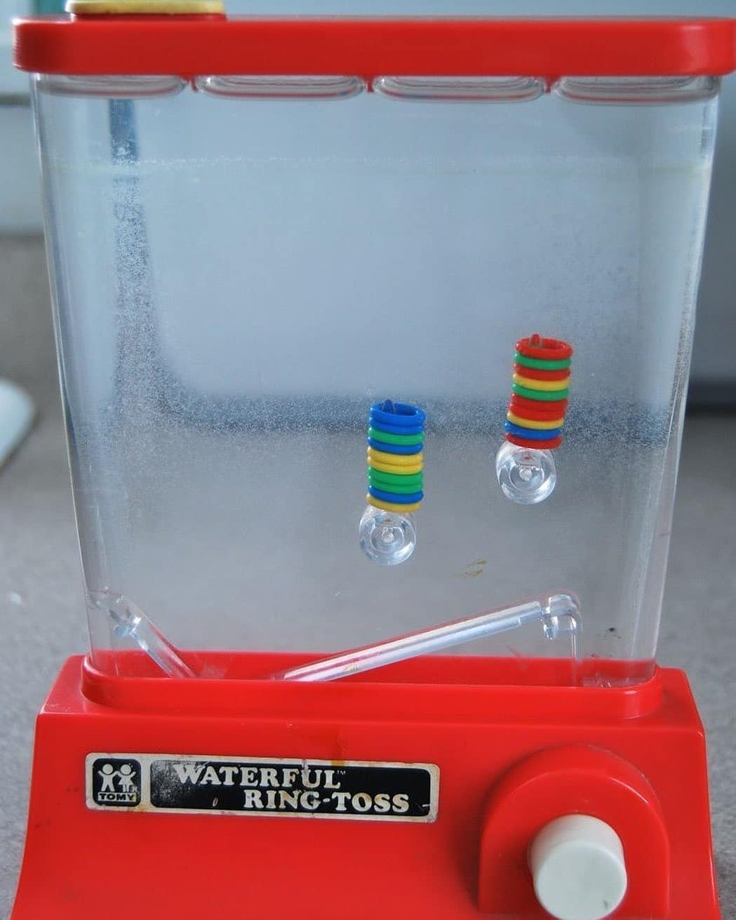 Juego de agua con anillas de colores y un botón rojo.