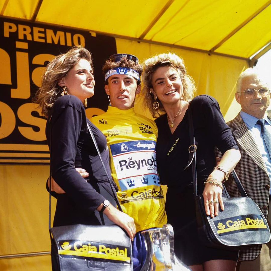 Ciclista con el maillot amarillo de la Vuelta en el podio, entre dos azafatas con bolsos de Caja Postal.