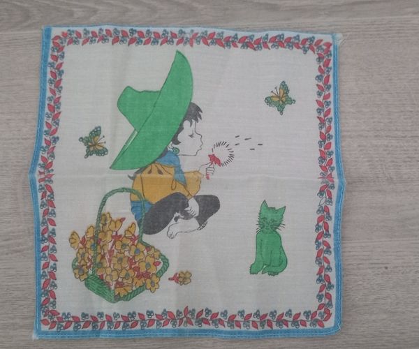 Pañuelo de tela con un niño con sombrero verde, un cesto de flores y un gato.