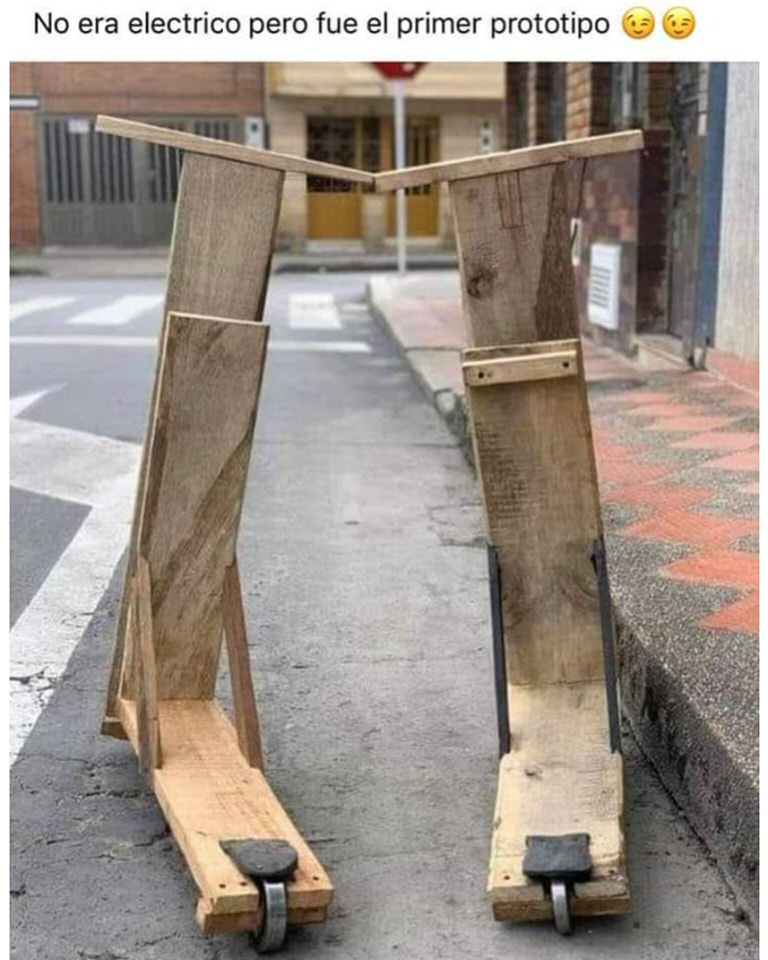 Dos patinetes hechos a mano con tablas de madera y ruedas pequeñas, apoyados en el asfalto junto a una acera.