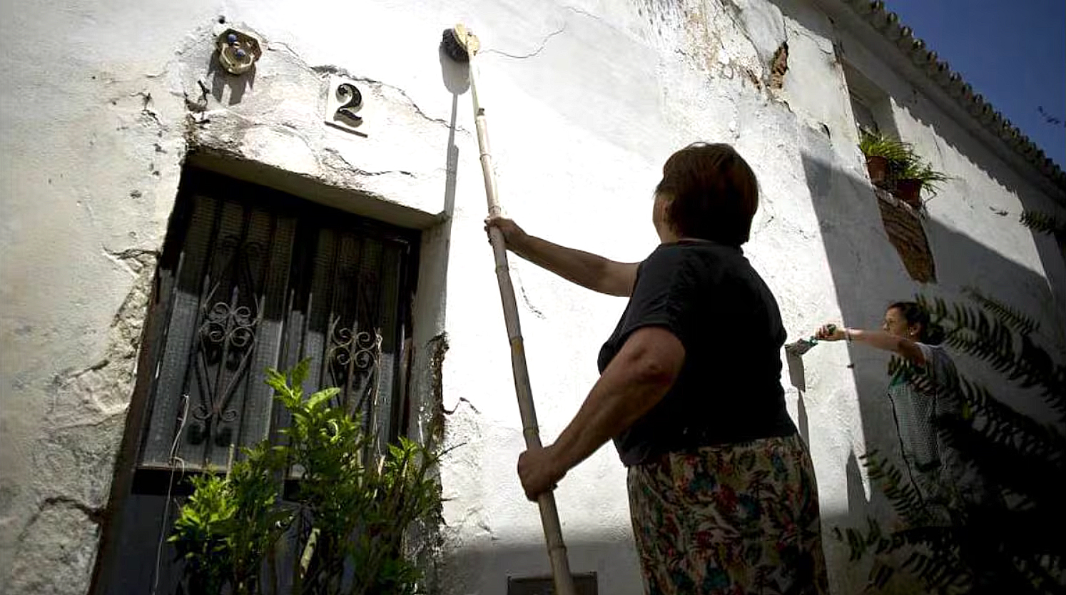 Mujer encalando la fachada de una casa con una escoba larga