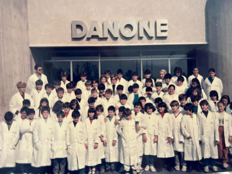 Grupo de escolares con batas blancas ante la entrada de una fábrica de Danone