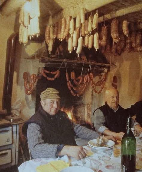 Dos hombres sentados a la mesa junto a chorizos colgados en una cocina de pueblo