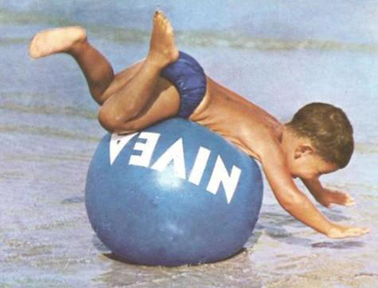 Niño en la playa tirándose sobre una pelota azul de Nivea.