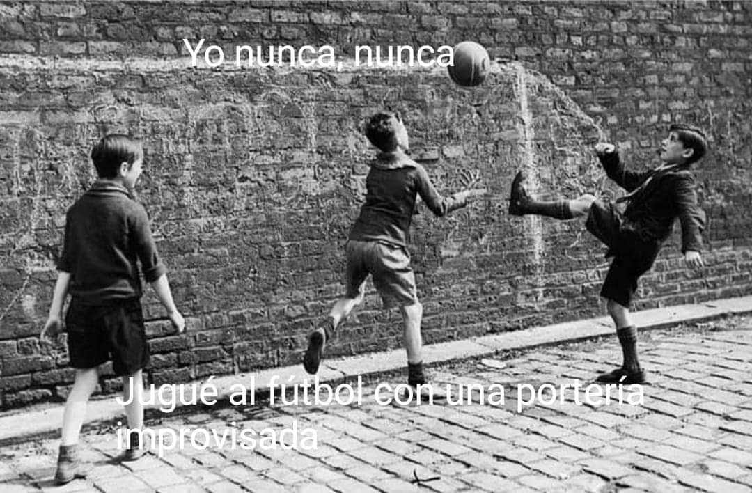 Foto en blanco y negro de tres niños jugando al fútbol contra una pared de ladrillo en una calle empedrada.