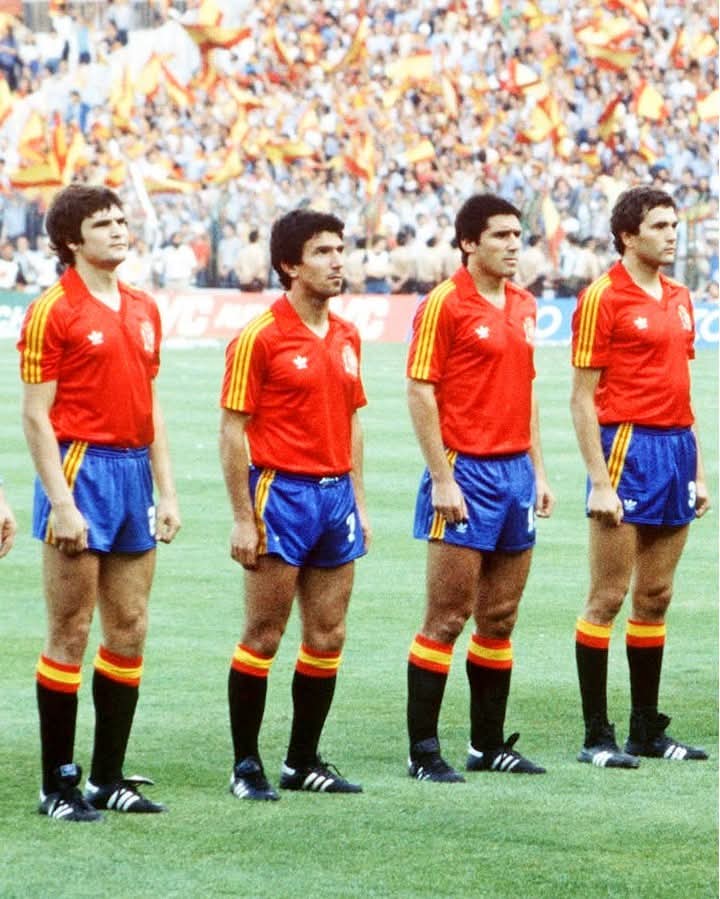 Cuatro futbolistas de la selección española en fila en el campo ante una grada llena de banderas.