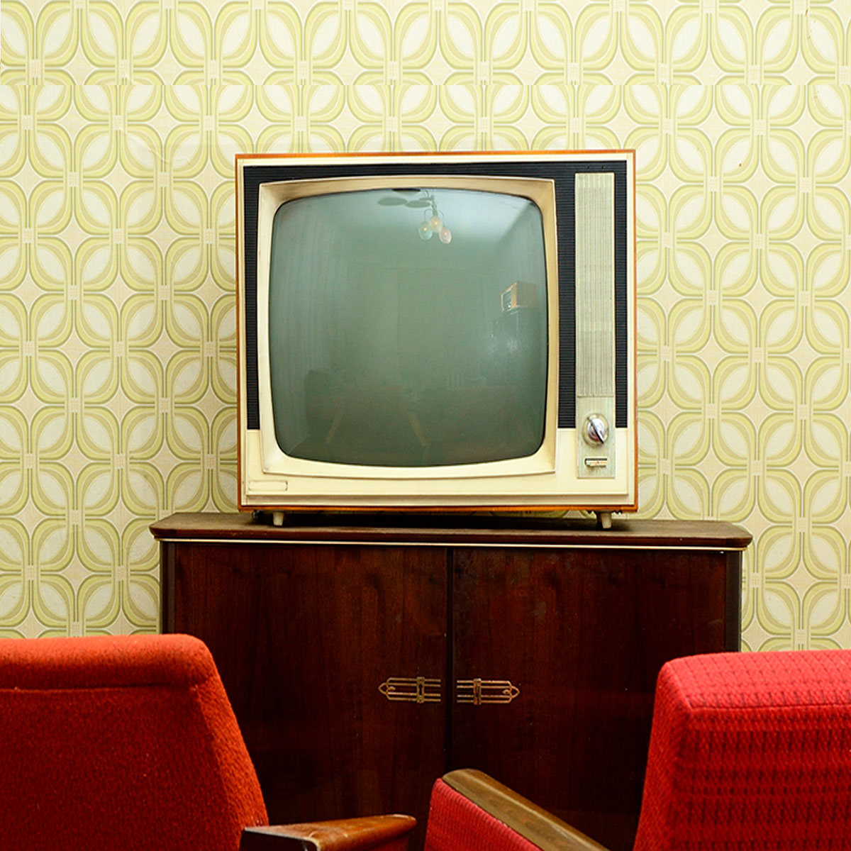 Televisor antiguo de color crema sobre un mueble de madera oscura, con papel pintado verde y dos sillones rojos.