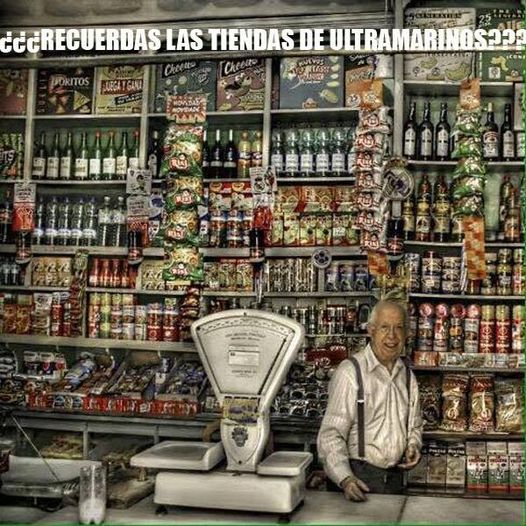 Tendero mayor detrás del mostrador de una tienda de ultramarinos llena de botellas, latas y bolsas de patatas colgadas.