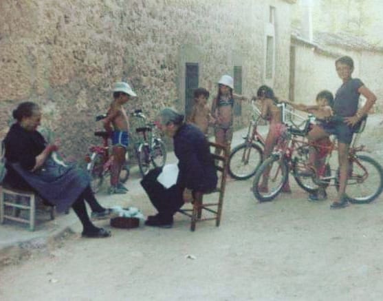 Niños con bicicletas junto a dos mujeres mayores sentadas en una calle de pueblo