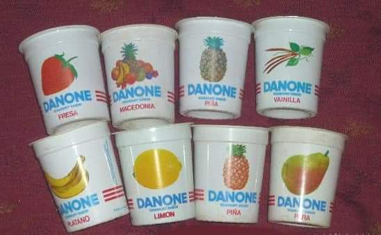 Ocho envases antiguos de yogur Danone con dibujos de frutas sobre un mantel rojo.