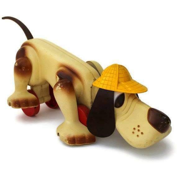 Perro de juguete de plástico color crema con orejas negras, gorro amarillo y ruedas rojas.