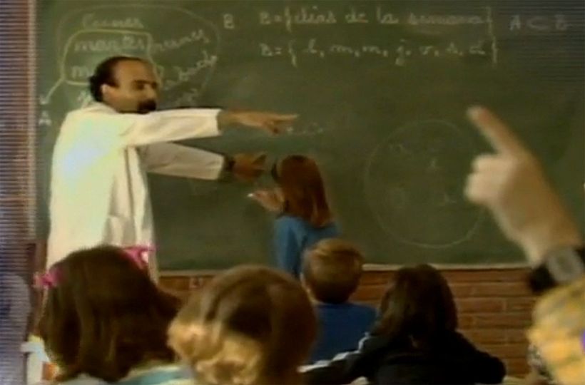 Profesor señalando la pizarra mientras los alumnos levantan la mano