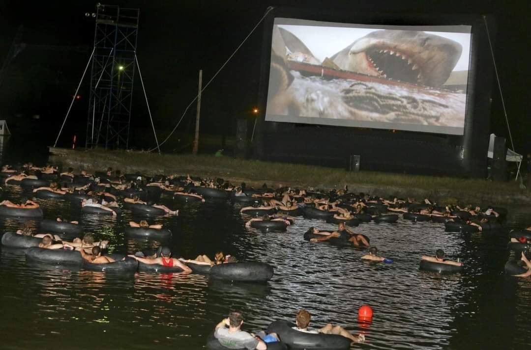 Gente en flotadores en un lago viendo Tiburón en una pantalla gigante.