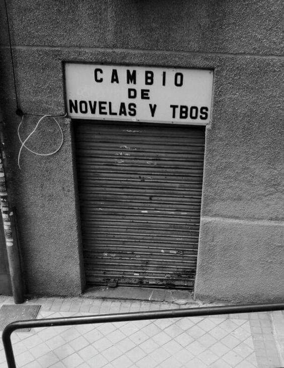 Local cerrado con persiana metálica y un cartel encima que dice Cambio de novelas y tebeos, en blanco y negro.